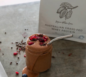 A Glass of mixed Chocolate cacao whey protein powder with rasberries and dark chocolate, with Australian Natural Protein Company Packaging of the Organic Cocoa Protein Powder flavour in the background