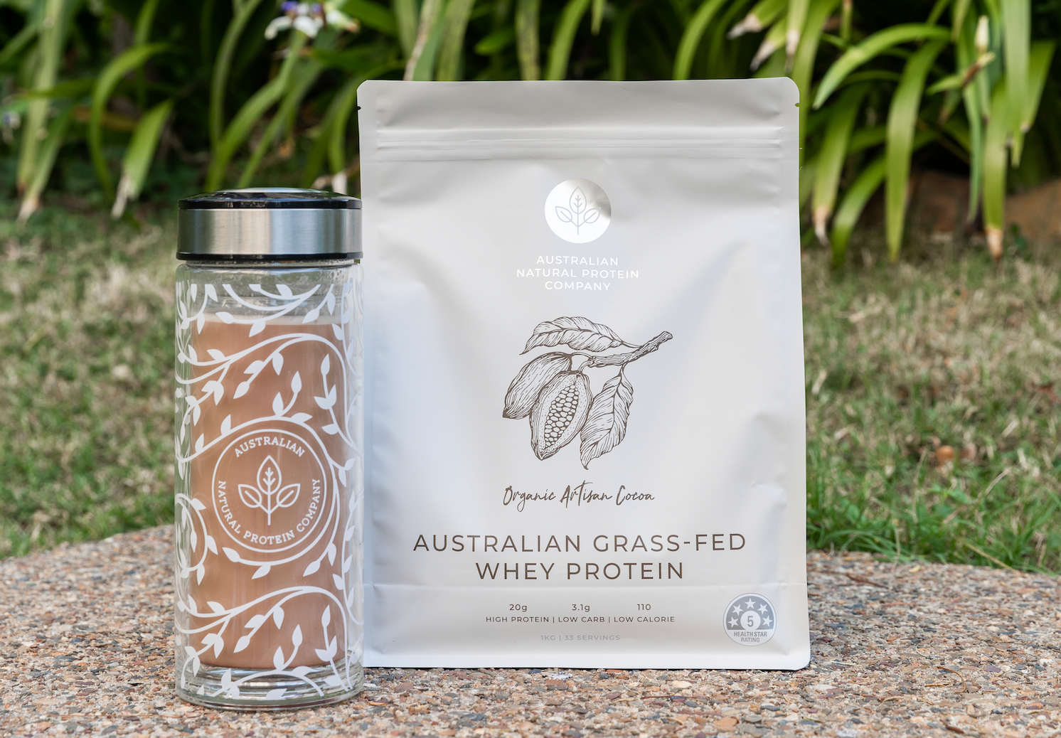 An ANPC Double Walled Glass Drink Bottle filled with mixed up protein shake using Chocolate cacao whey protein powder, next to a packet of Australian Natural Protein Company Packaging of the Organic Cocoa Protein Powder flavour