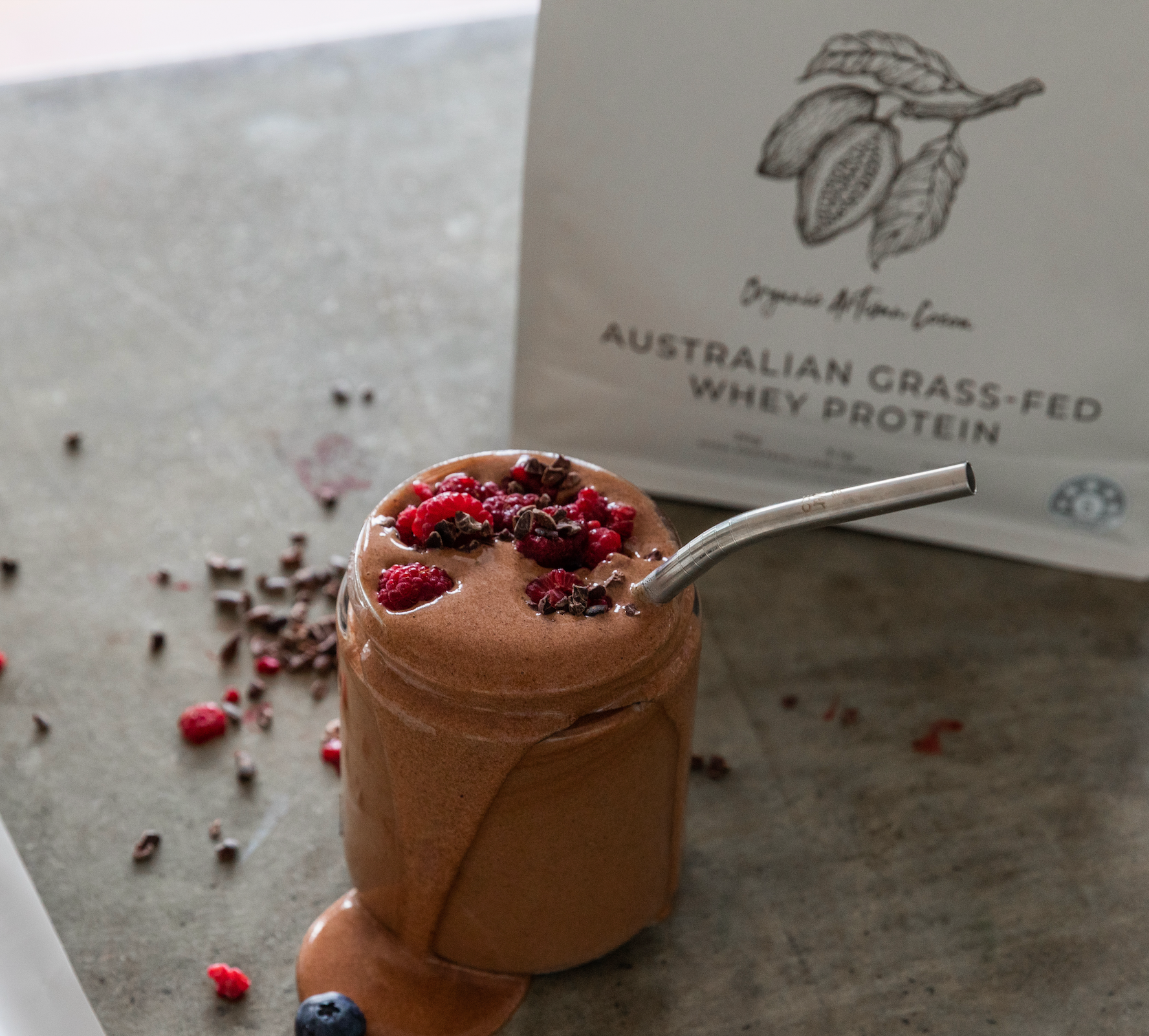A Glass of mixed Chocolate cacao whey protein powder with rasberries and dark chocolate, with Australian Natural Protein Company Packaging of the Organic Cocoa Protein Powder flavour in the background